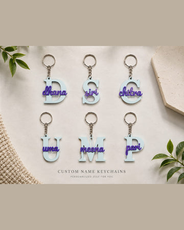 Custom name alphabet keychains laid flat: light blue initial letters D, S, C, U, M and P with purple script names and silver key rings